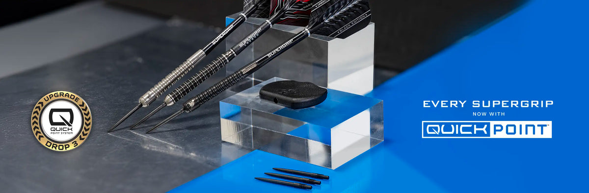 A promotional graphic for Quick Point darts, showing several black and silver SuperGrip barrels with flights inserted in clear acrylic display blocks on a dark tabletop. The right side has a blue graphic panel with the text 'EVERY SUPERGRIP TYPE A/1 QUICK POINT' and a circular 'Program of Quality' badge on the left.